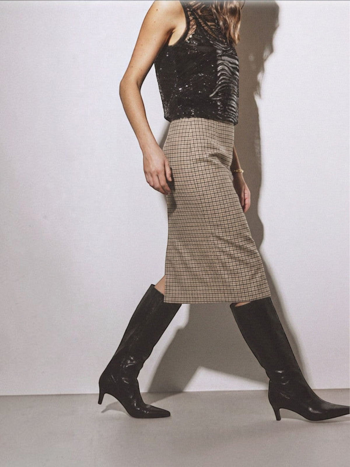 Clueless Paris midi skirt in neutral plaid with a side slit, worn with black knee-high boots and a sequin top.