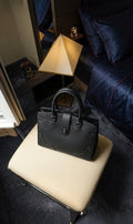 Sac à main Clueless Paris en cuir grainé noir avec fermoir métallique, posé sur un pouf dans une chambre élégante.